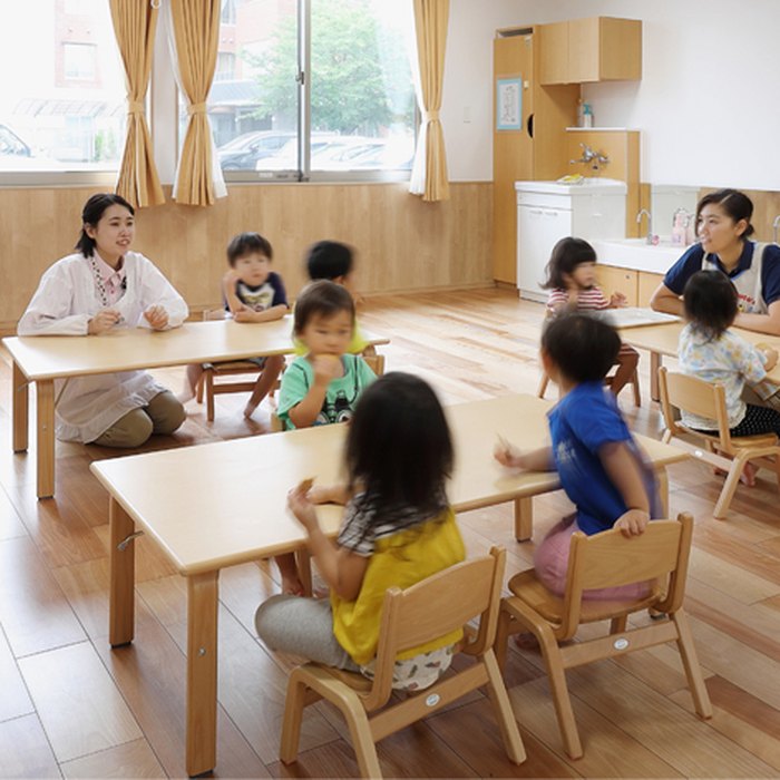 保育室で机を囲み、保育士と一緒に過ごす子どもたち