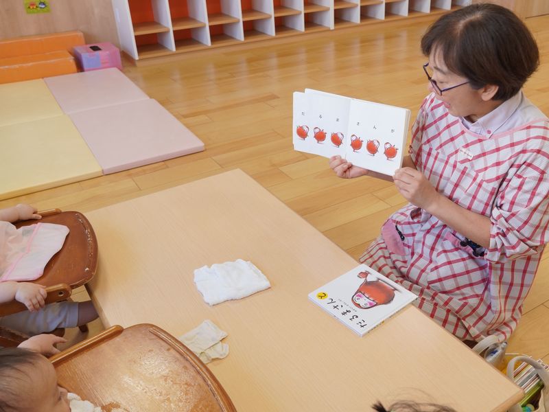 0歳児クラスの保育室で、保育士が絵本を見せながら朝の会をしている様子