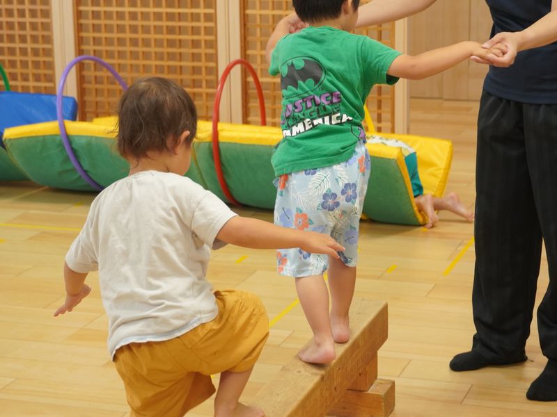 遊戯室で平均台を渡って遊ぶ1・2歳児クラスの子どもたち